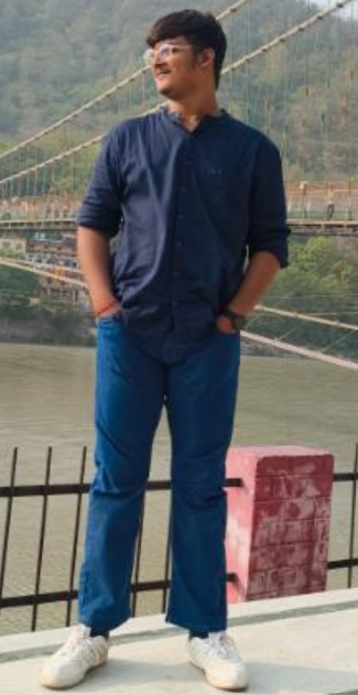 A young man enjoying the beautiful outside scenery while standing boldly next to a river and suspension bridge in blue slacks and a navy blue blouse.