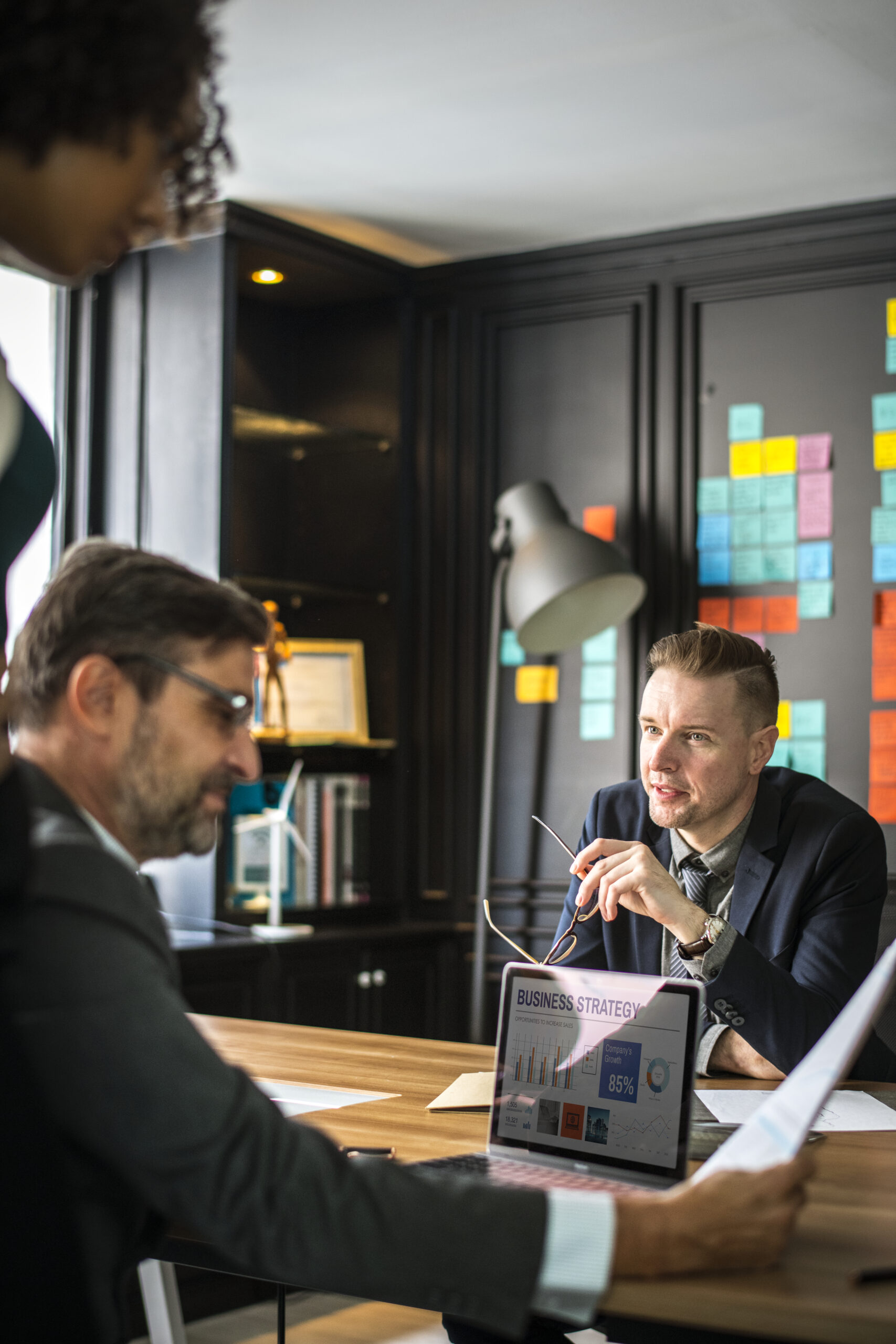 With charts and post-it notes in the backdrop, three business executives are in a meeting discussing the company's growth strategy.
