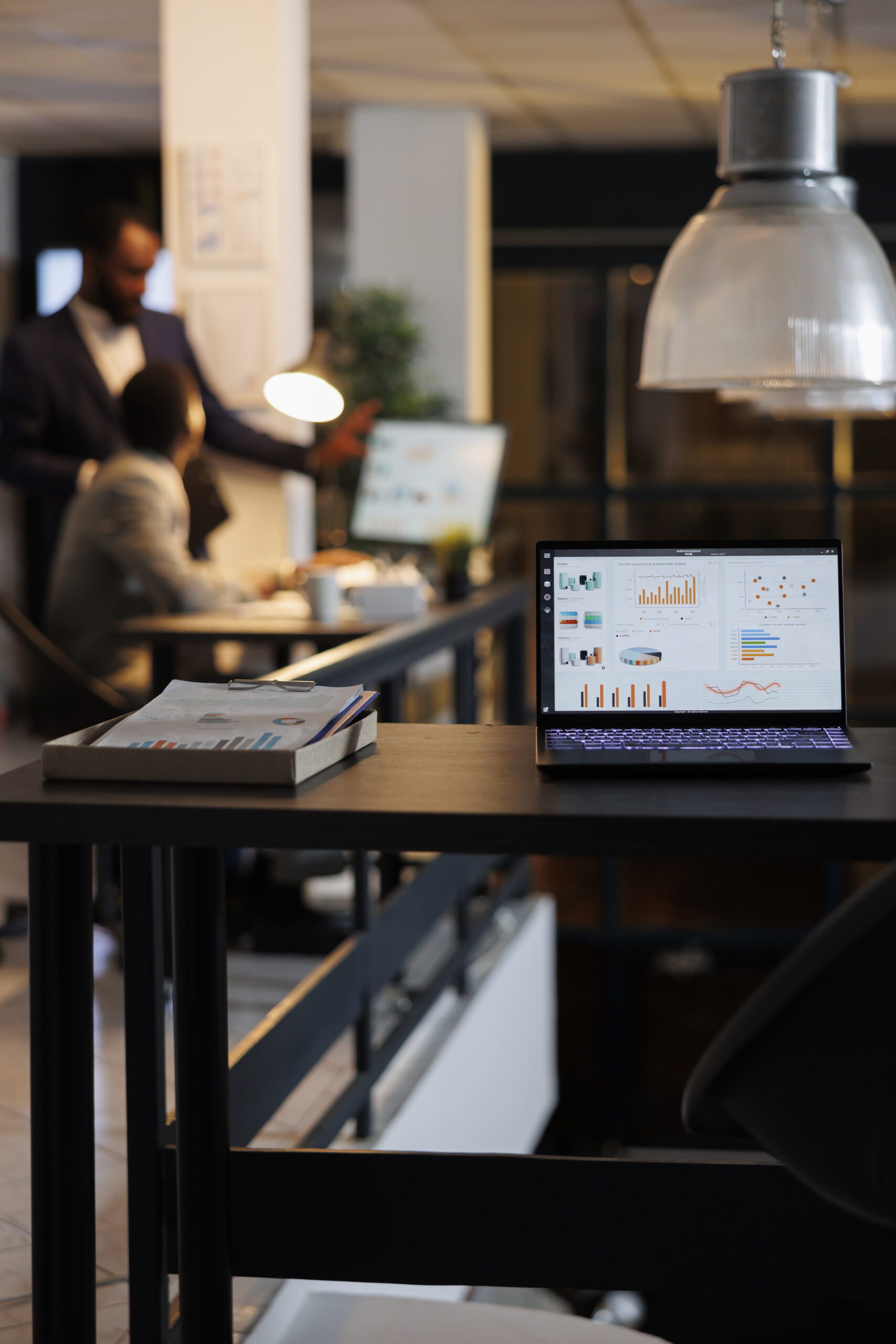 Coworkers discussing business strategy in the background of an open laptop displaying financial graphs and charts on a desk in a contemporary startup workplace.