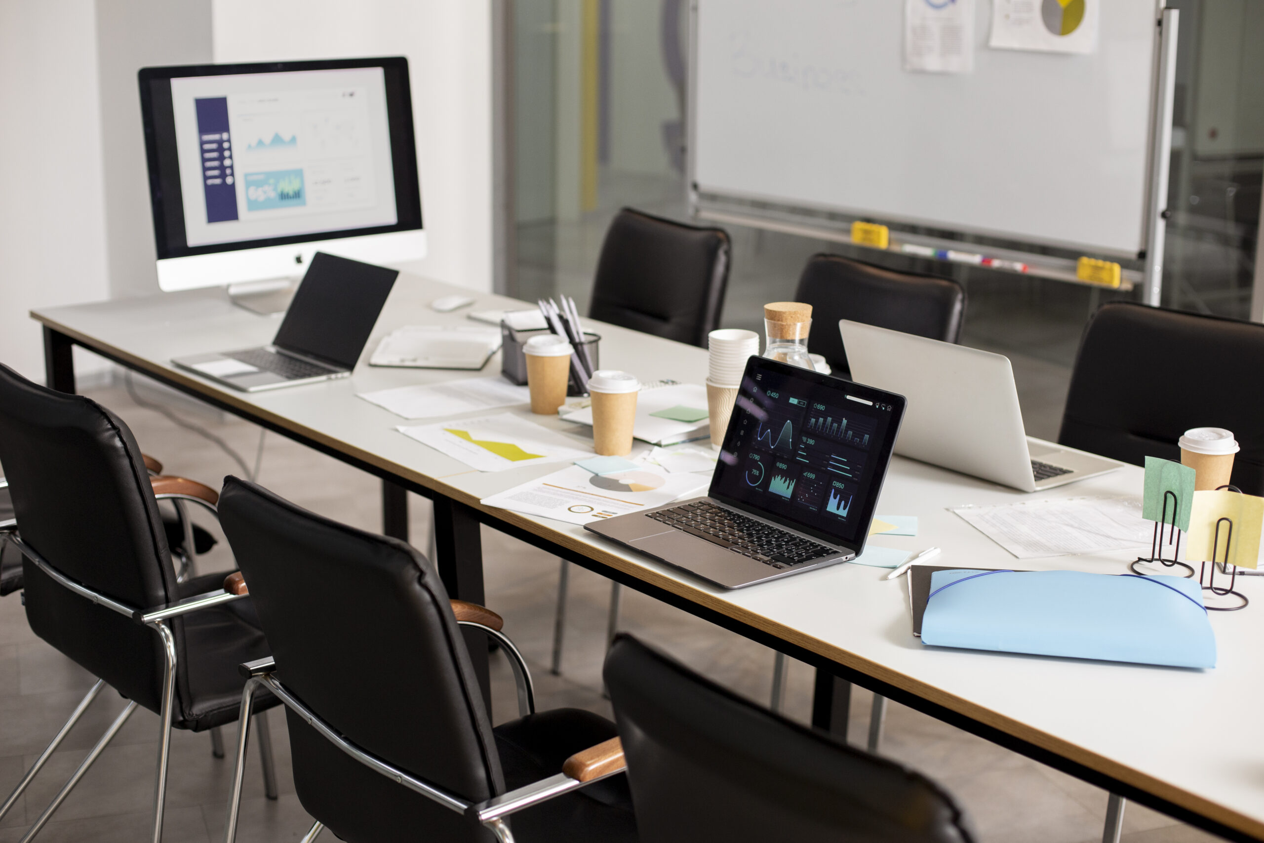 For a marketing strategy meeting, a contemporary office conference room is prepared with laptops, charts, coffee cups, and business analytics dashboards on displays.